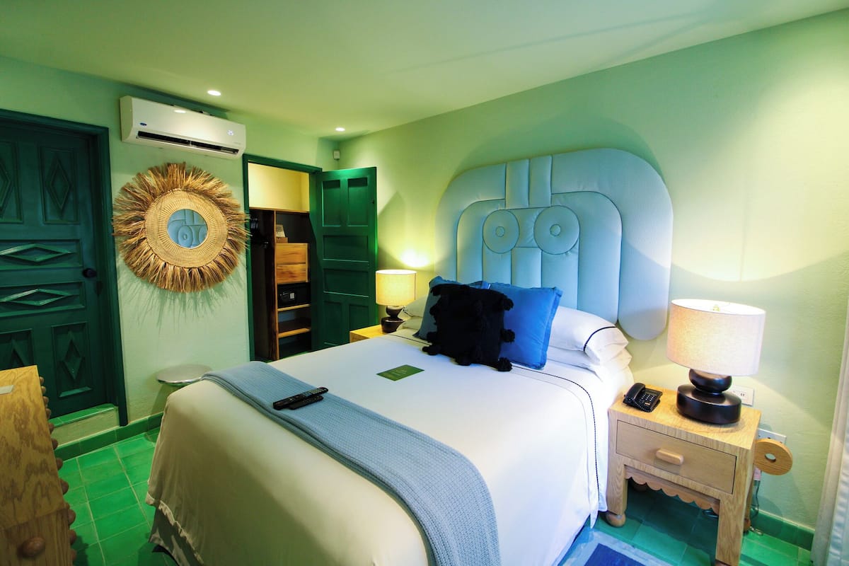 A hotel room with a bed, bedside tables, a lamp, a wall-mounted air conditioner, and a decorative wall hanging.