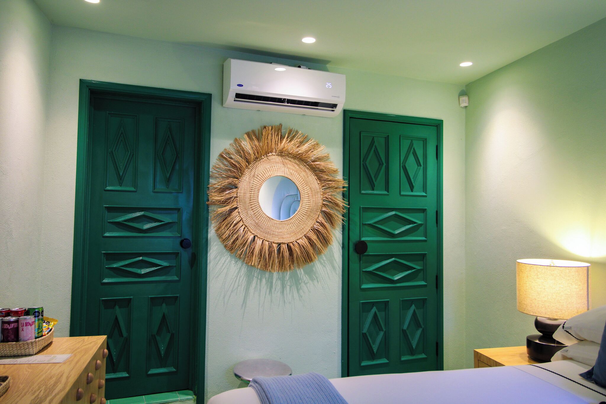 Habitación con dos puertas verdes, aire acondicionado de pared, espejo tejido, lámpara de noche y una pequeña mesa con objetos.