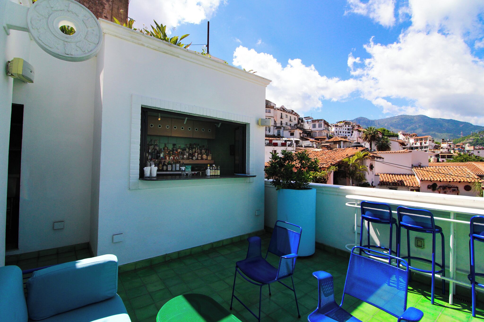 A rooftop terrace with a bar, seating, and a view of a town with buildings and mountains.