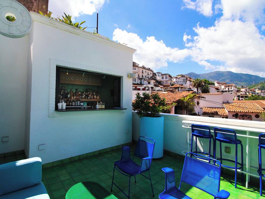 A rooftop terrace with a bar, seating, and a view of a town with buildings and mountains.