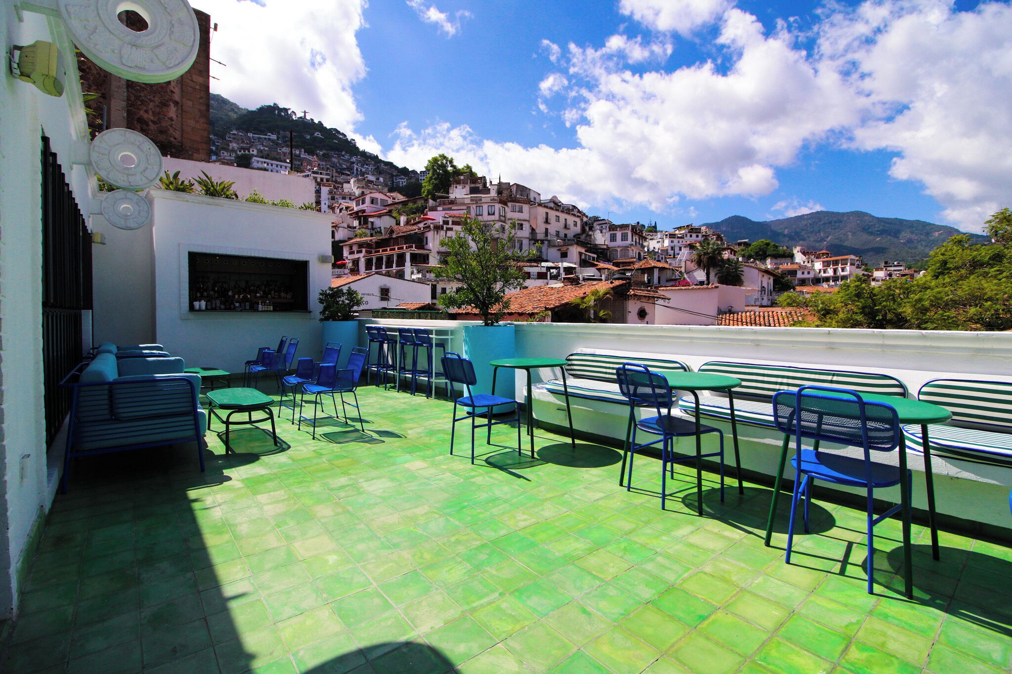 A rooftop terrace with green flooring, blue chairs, and tables, overlooking a hillside town with buildings and mountains.