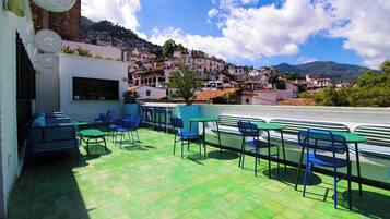 A rooftop terrace with green flooring, blue chairs, and tables, overlooking a hillside town with buildings and mountains.