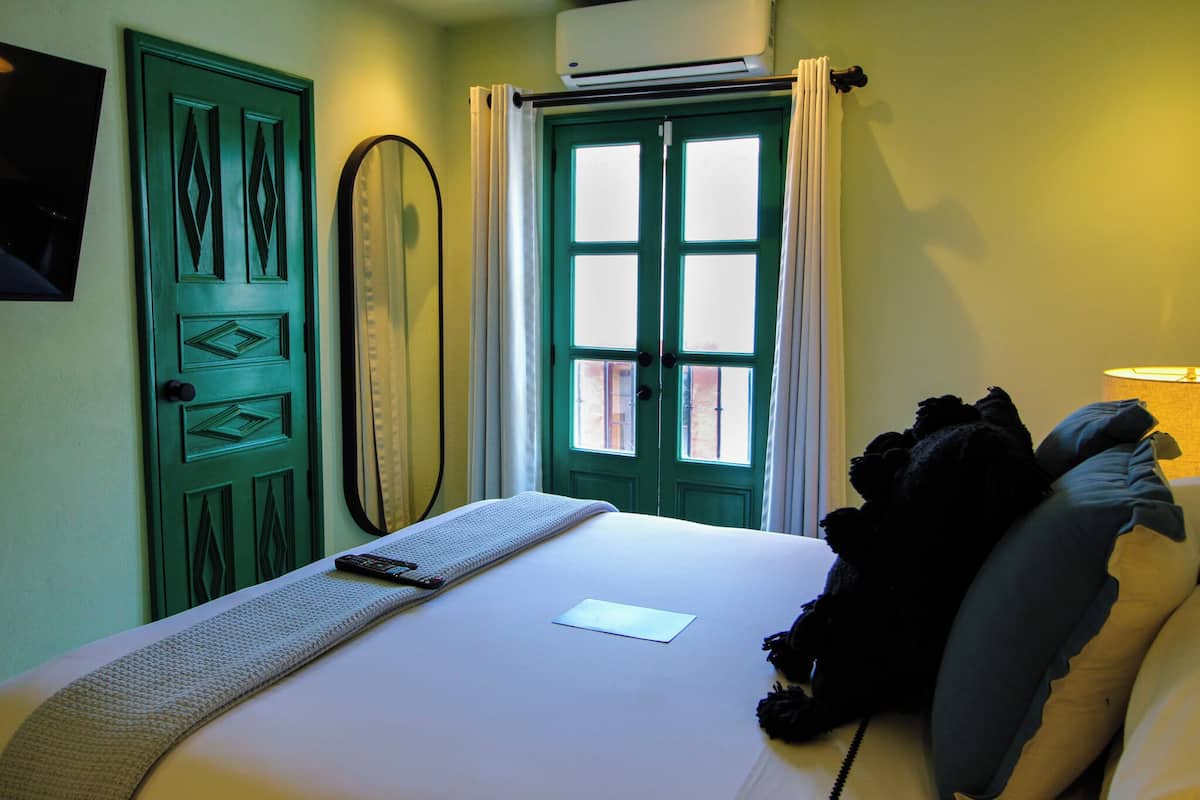 A hotel room with a bed, a window with curtains, a door with a decorative design, and an air conditioning unit.