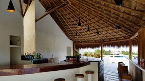 Un área de bar con techo de paja, paredes blancas y piso de baldosas. Hay taburetes de bar y una barra con botellas y vasos.