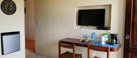 Una habitación con una mesa de madera, dos sillas, un televisor montado en la pared, una cafetera y un dispensador de agua.