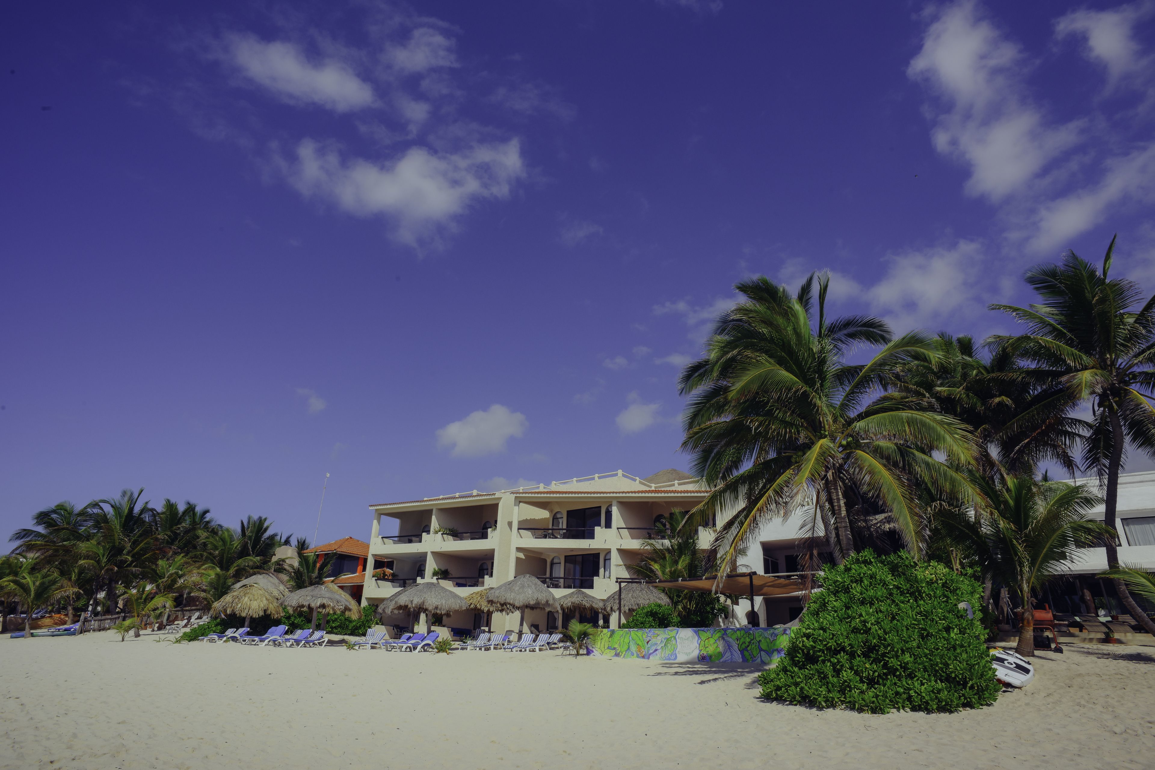 A beachfront hotel with multiple balconies, palm trees, and thatched umbrellas.