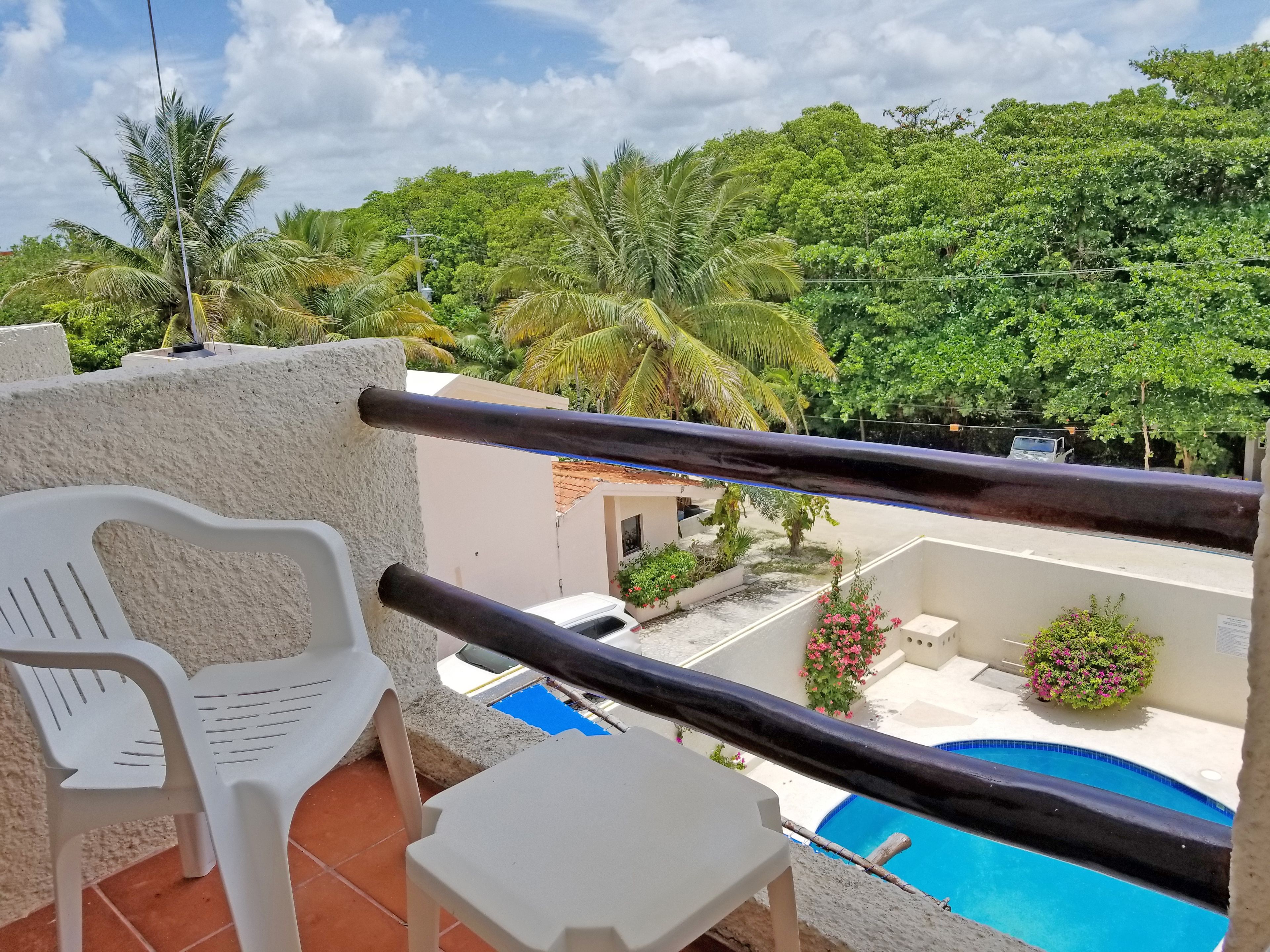 A balcony with a pool, white plastic chairs, and a view of a lush green landscape.