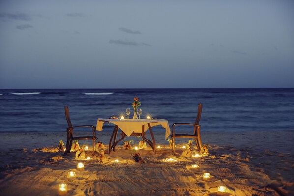 Montaje para cena en la playa con mesa, sillas, velas y vista al océano.