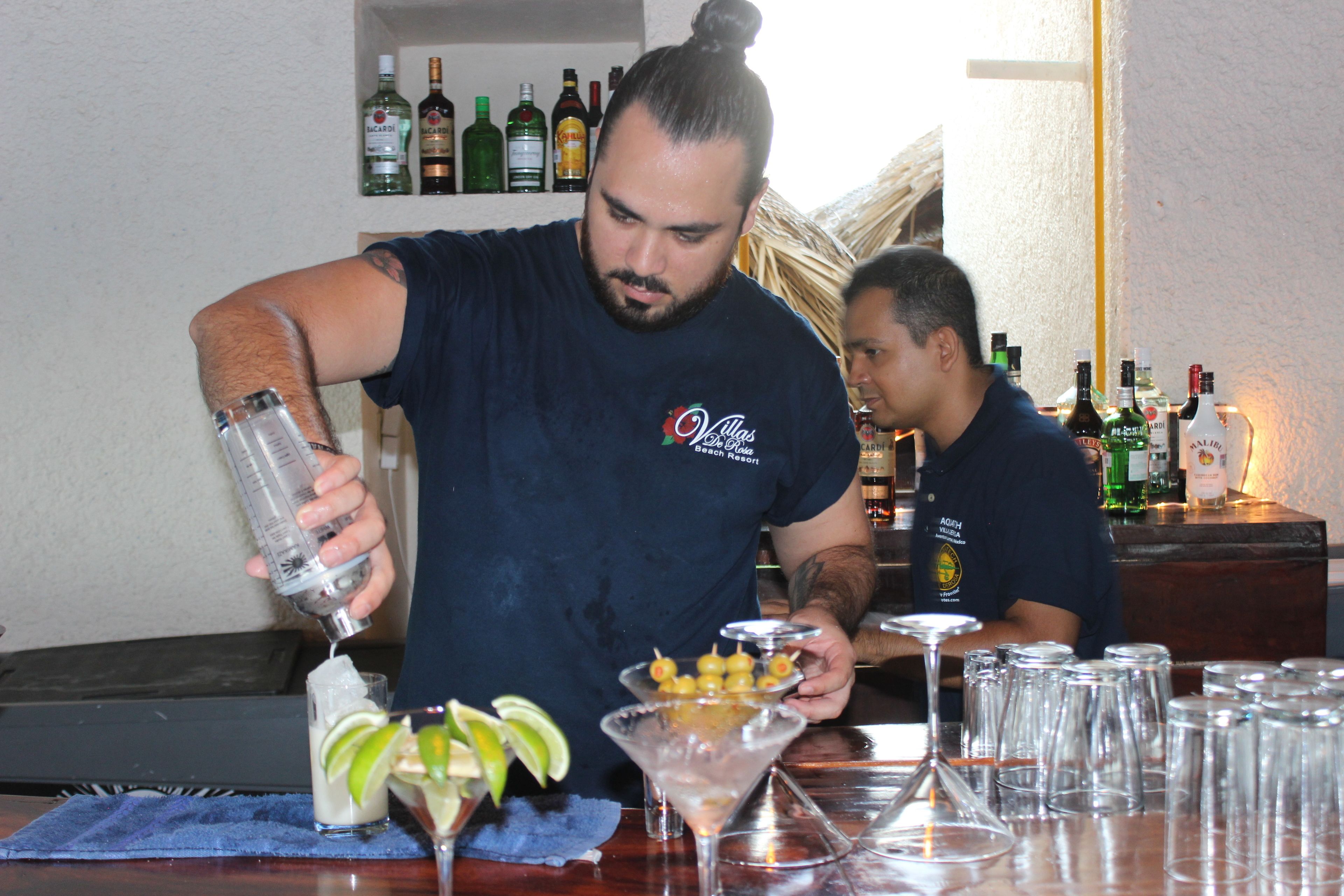 Un barman preparando una bebida en una barra, con varias botellas y vasos al fondo.