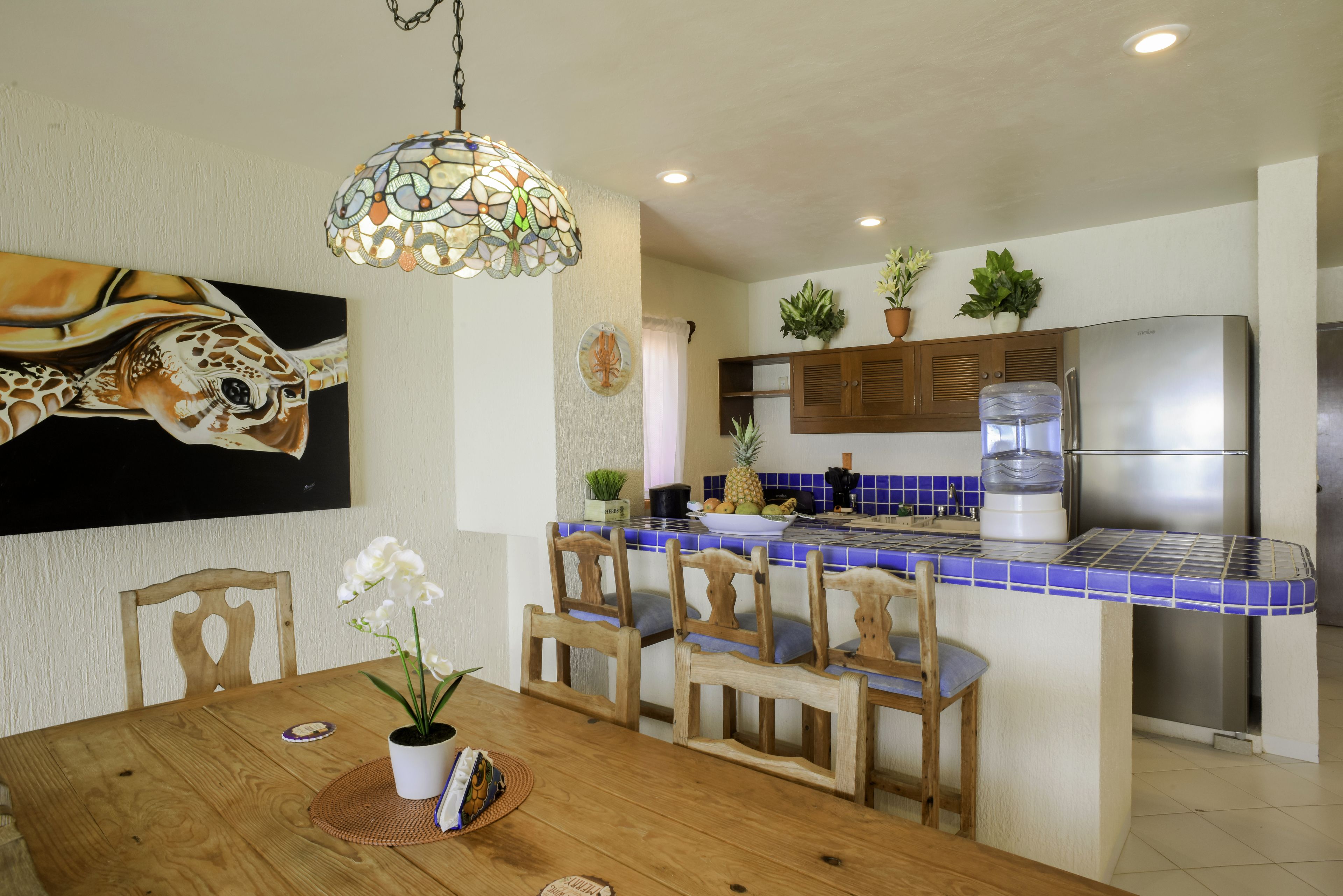 A kitchen with a blue-tiled backsplash, wooden chairs, a dining table with a white orchid, and a refrigerator.