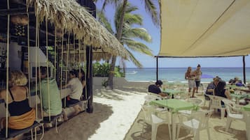 A beachside restaurant with outdoor seating, palm trees, and a view of the ocean.