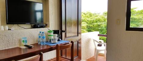 A room with a wooden table, chairs, a television mounted on the wall, a coffee maker, and a view of greenery outside.