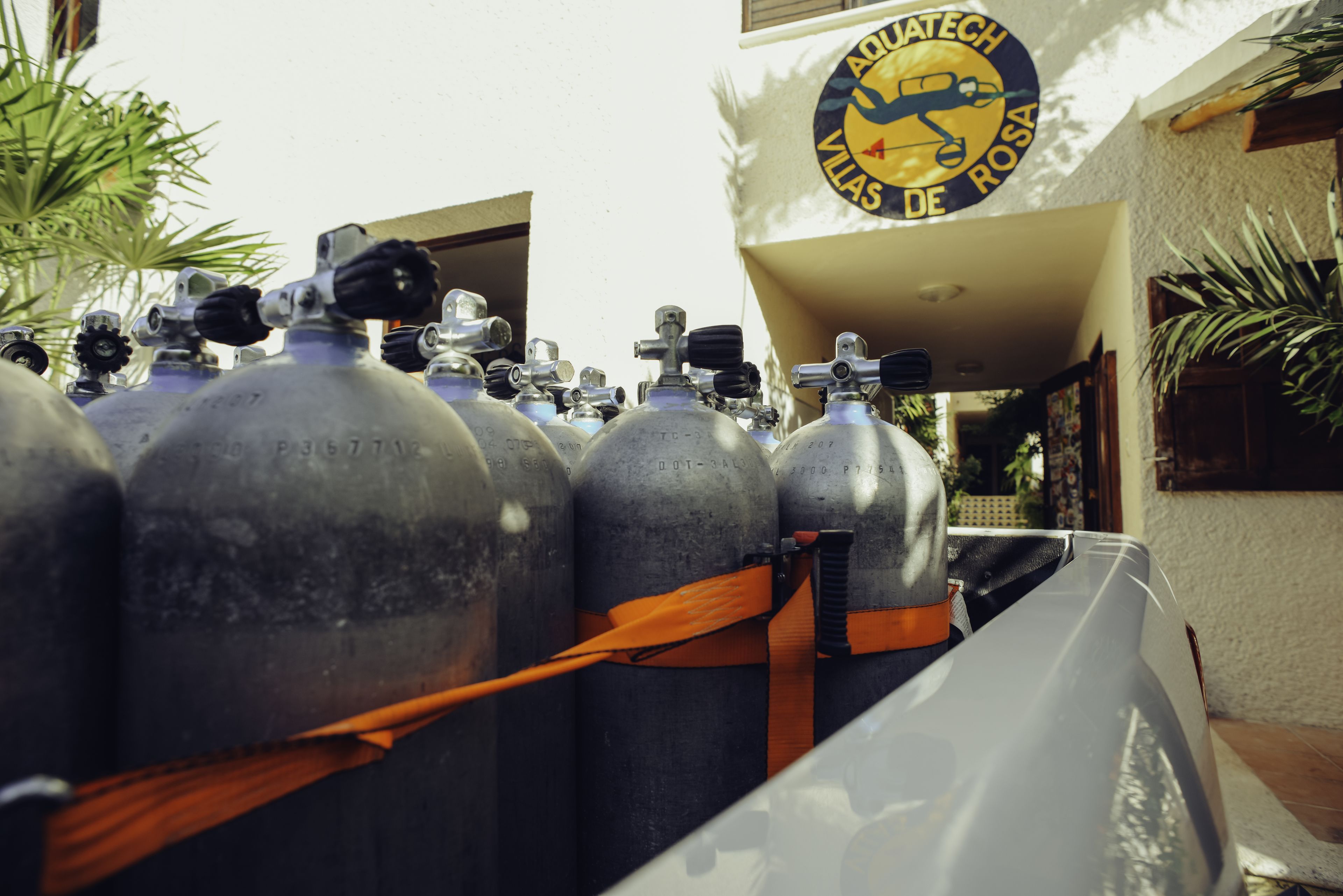 A row of large, metallic oxygen tanks secured with straps, with a building and a logo in the background.