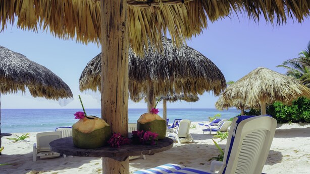 Escena de playa con sombrillas de paja, sillas de descanso y una mesa con cocos y flores.
