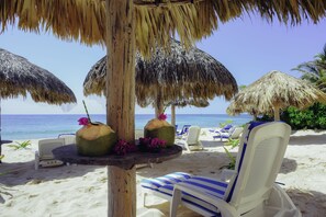 Escena de playa con sombrillas de paja, sillas de descanso y una mesa con cocos y flores.