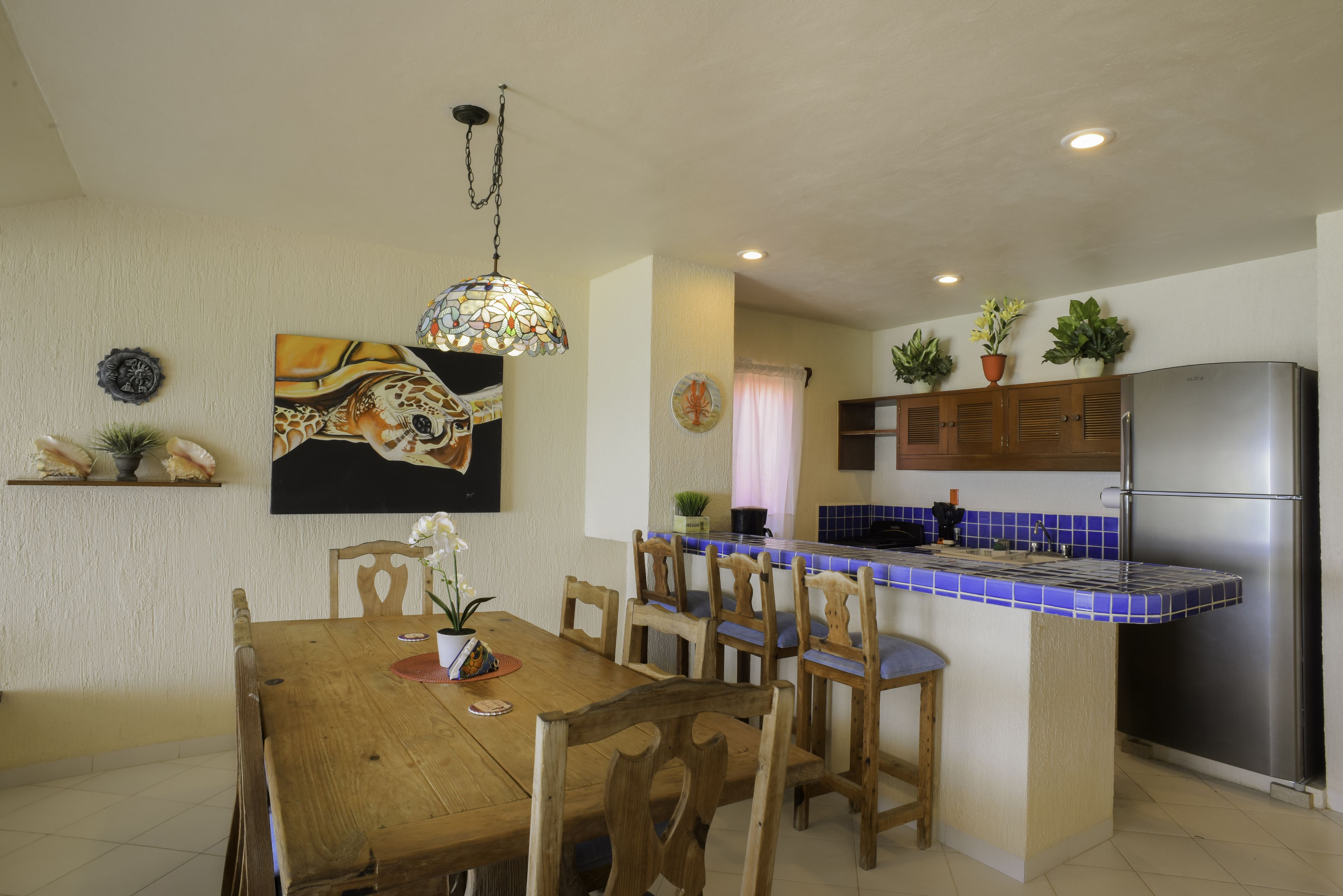A dining area with a wooden table and chairs, a hanging pendant light, and a kitchen with a stainless steel refrigerator and blue-tiled backsplash.