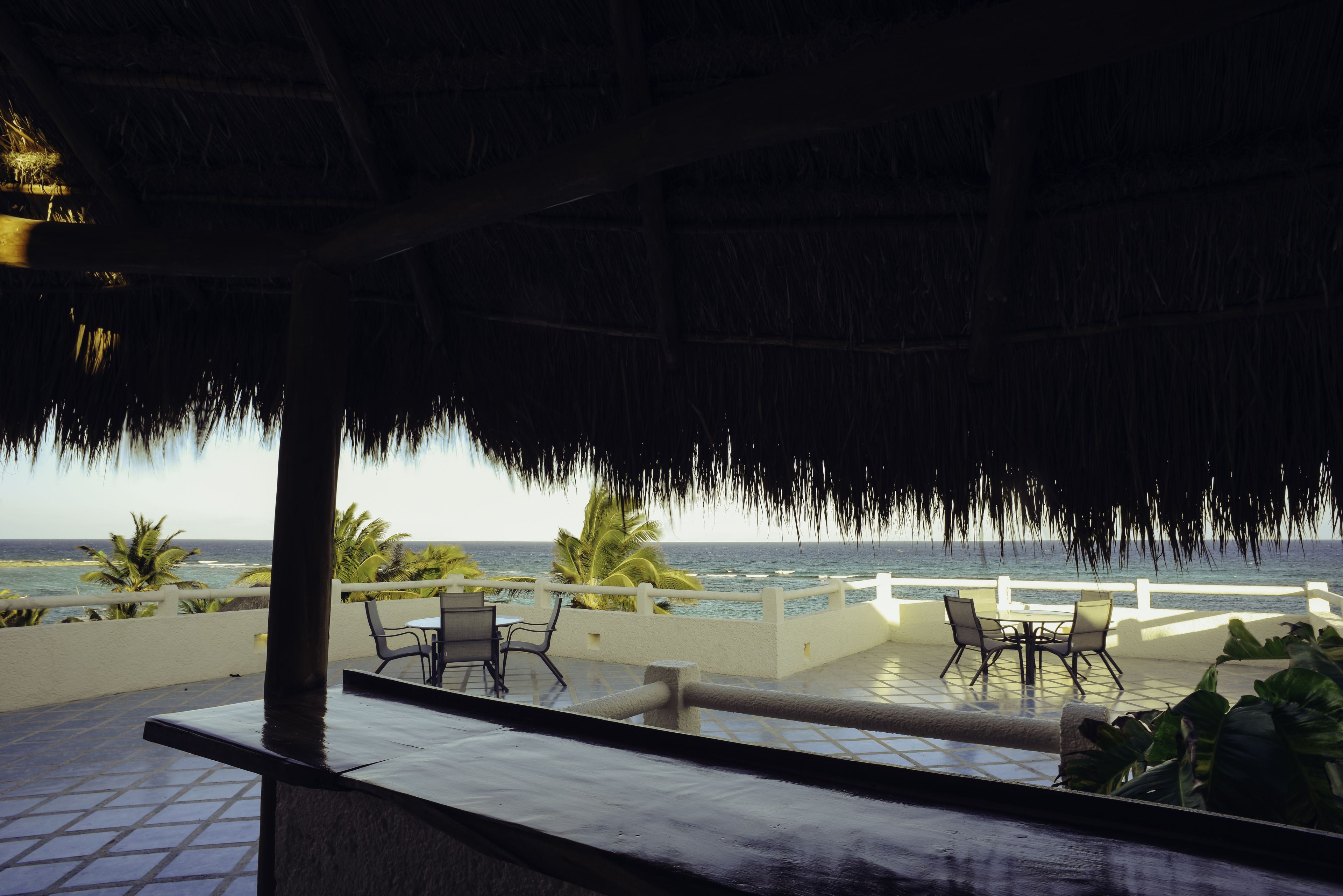 A covered outdoor seating area with a view of the ocean, featuring a thatched roof, chairs, and a table.