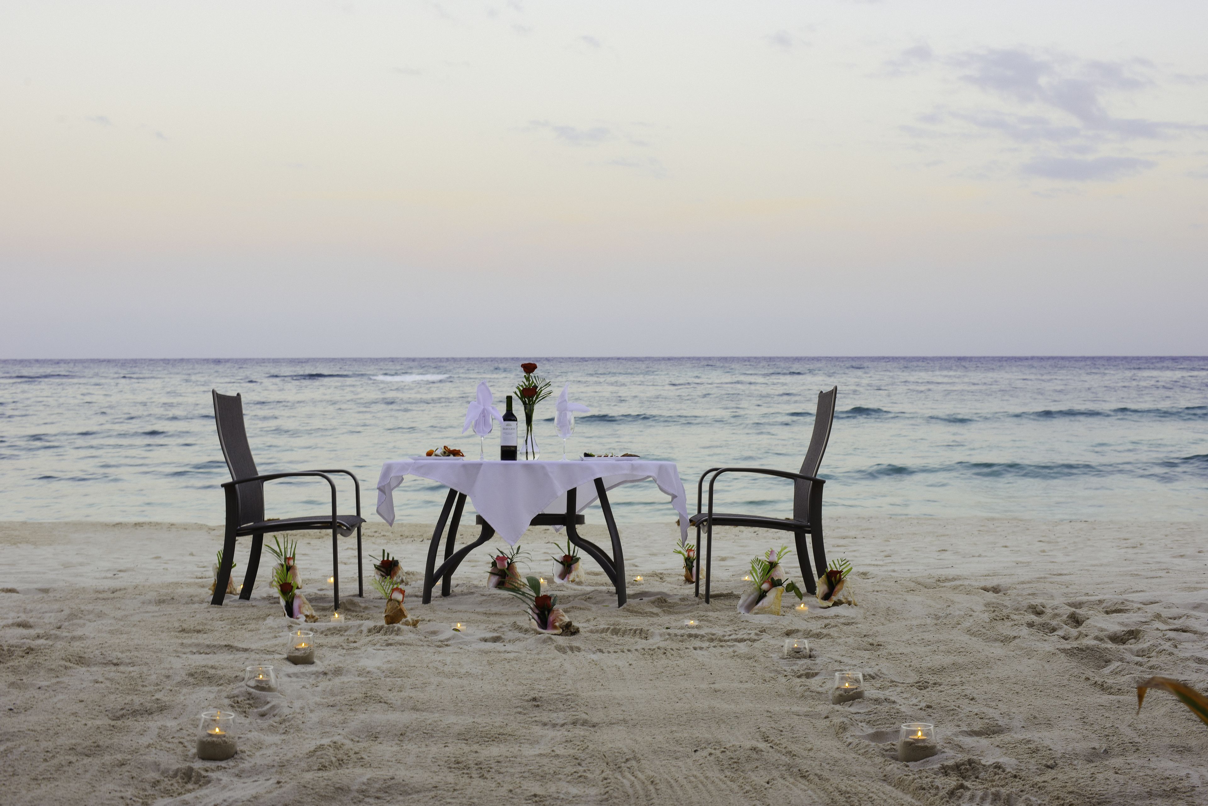 A beachside dining setup with a table and chairs, candles, and decorative plants.