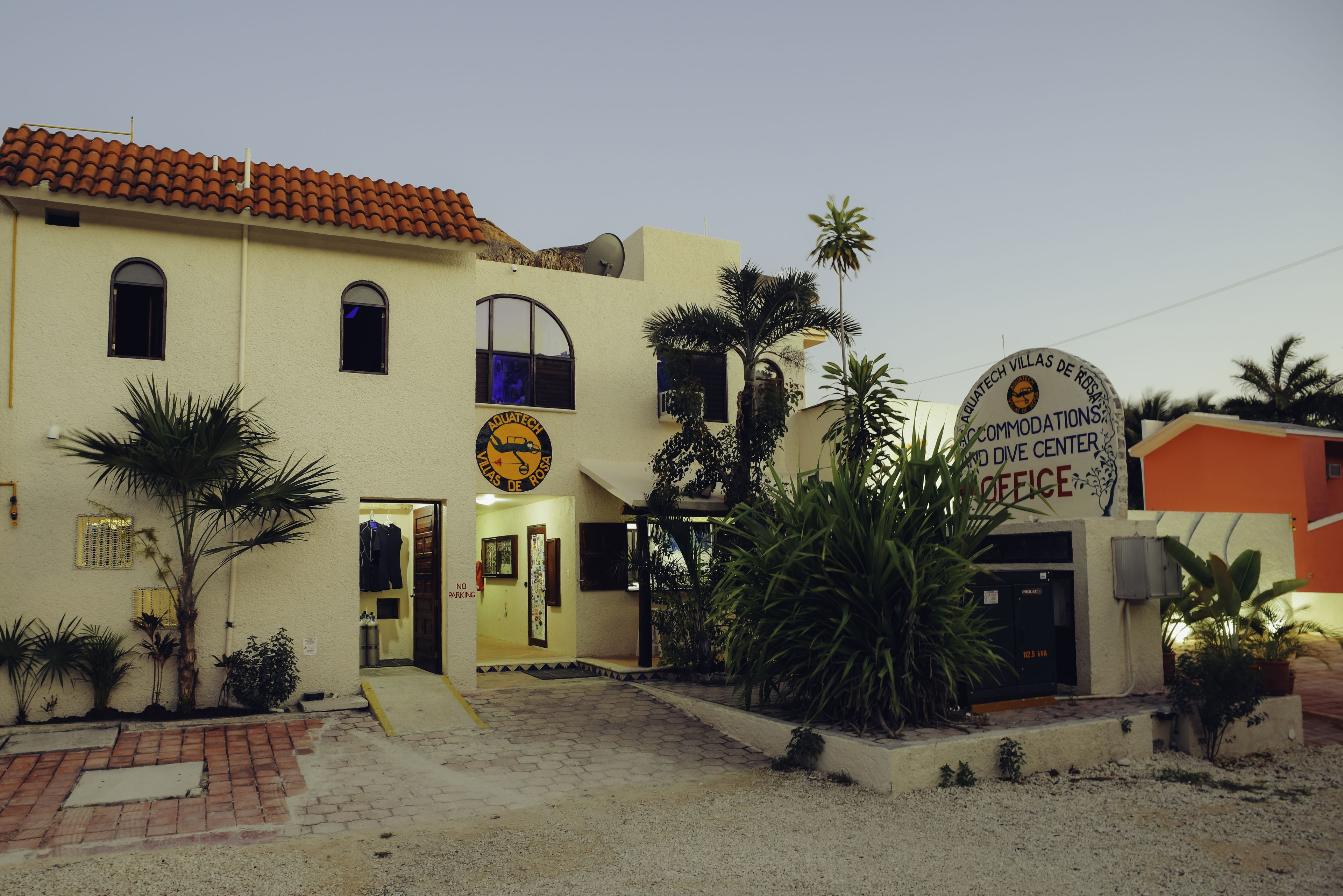 A building with a sign for Aquatech Villas de Rosario Dive Center and an office. There are palm trees and a brick pathway leading to the entrance.
