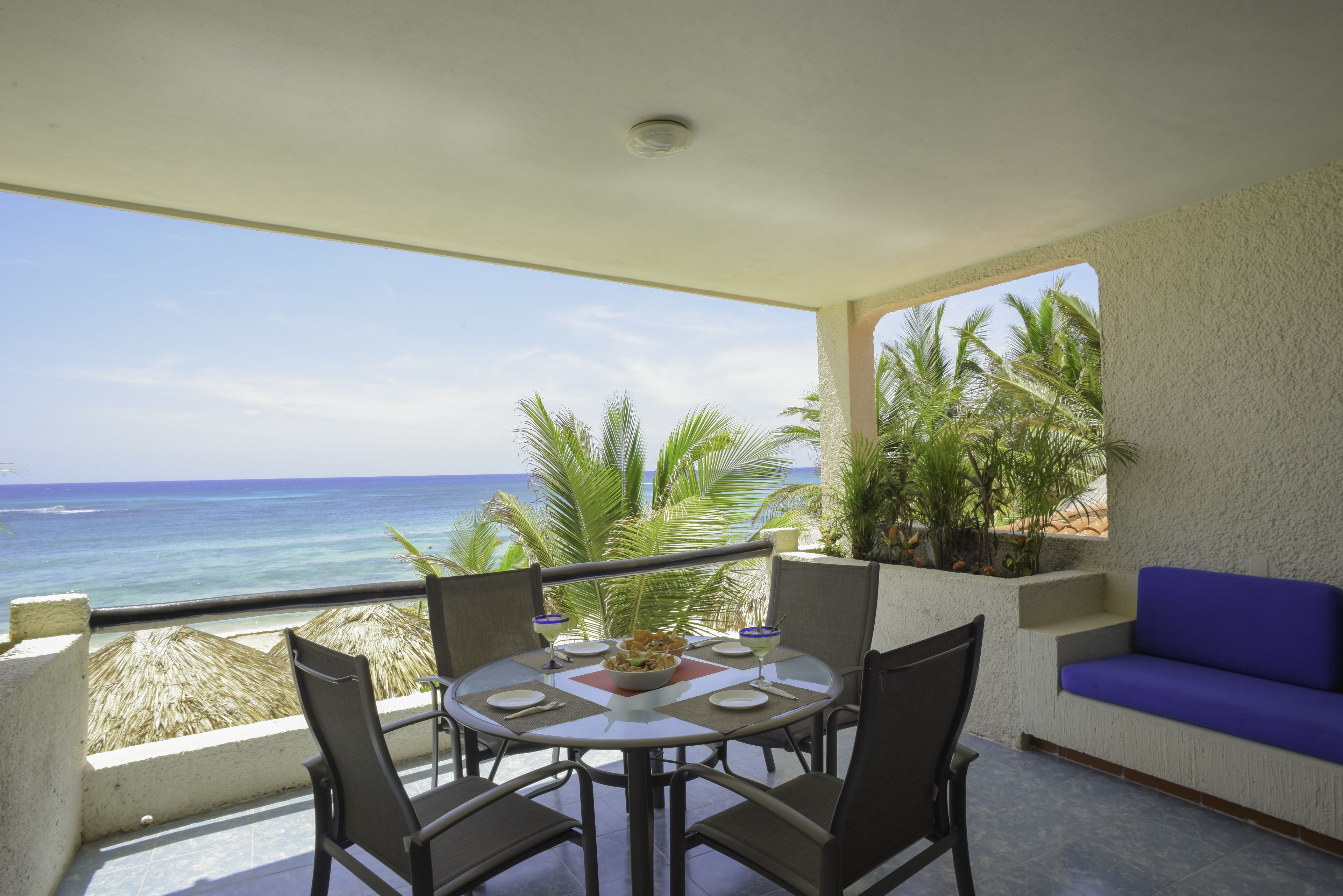 A balcony with a table and chairs, overlooking the ocean with palm trees.