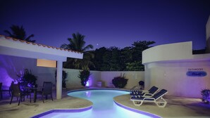 Zona de piscina con sillas de descanso, una mesa y un letrero que indica un baño.