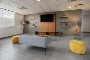 A modern waiting area with a grey sofa, yellow ottoman, a flat-screen TV mounted on a wooden panel, and shelves with small plants and decorative items.