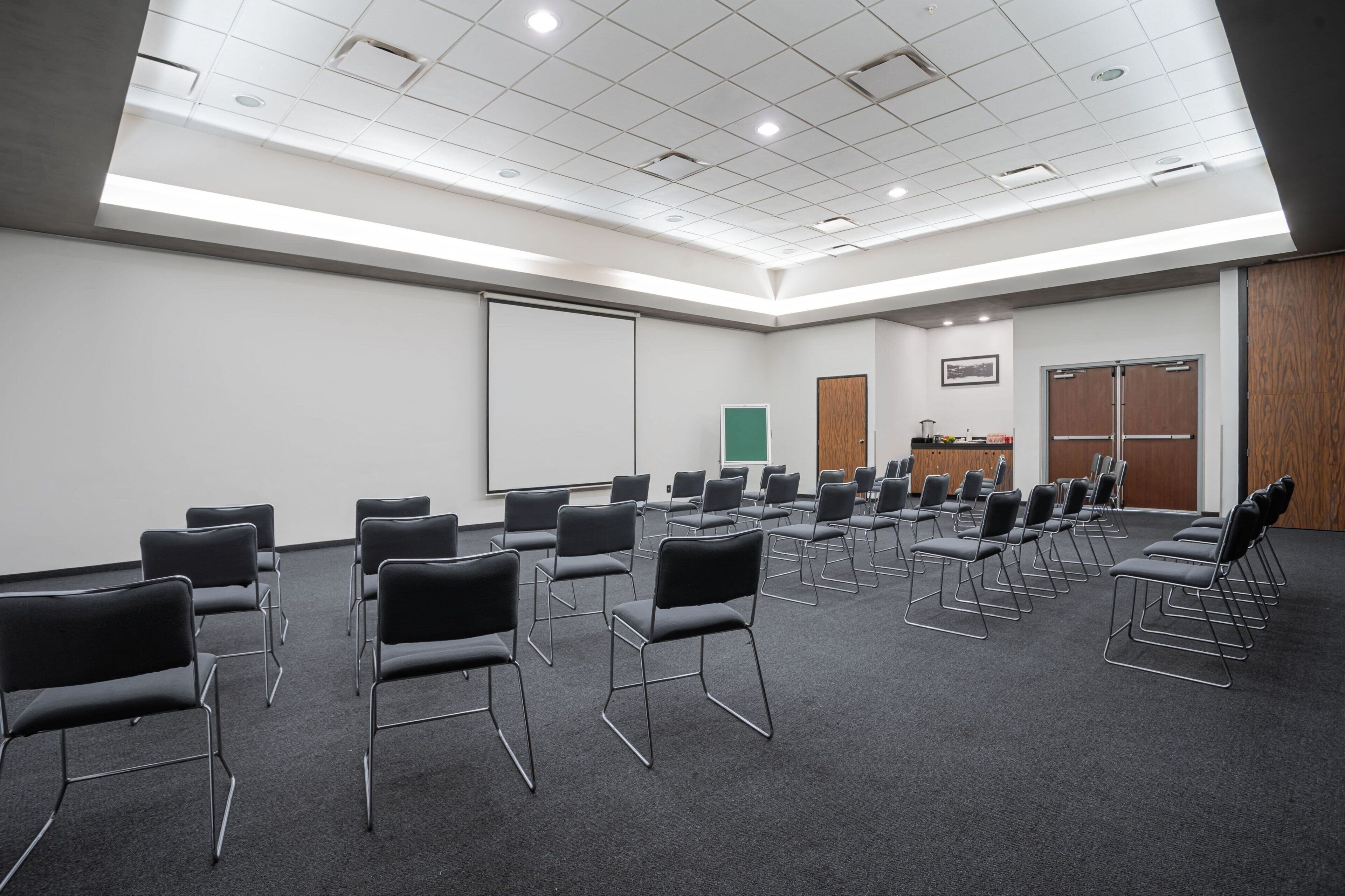 Un salón de conferencias con filas de sillas negras, una pizarra y una pantalla para proyector.