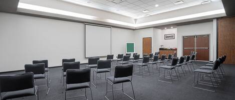 Un salón de conferencias con filas de sillas negras, una pizarra y una pantalla para proyector.