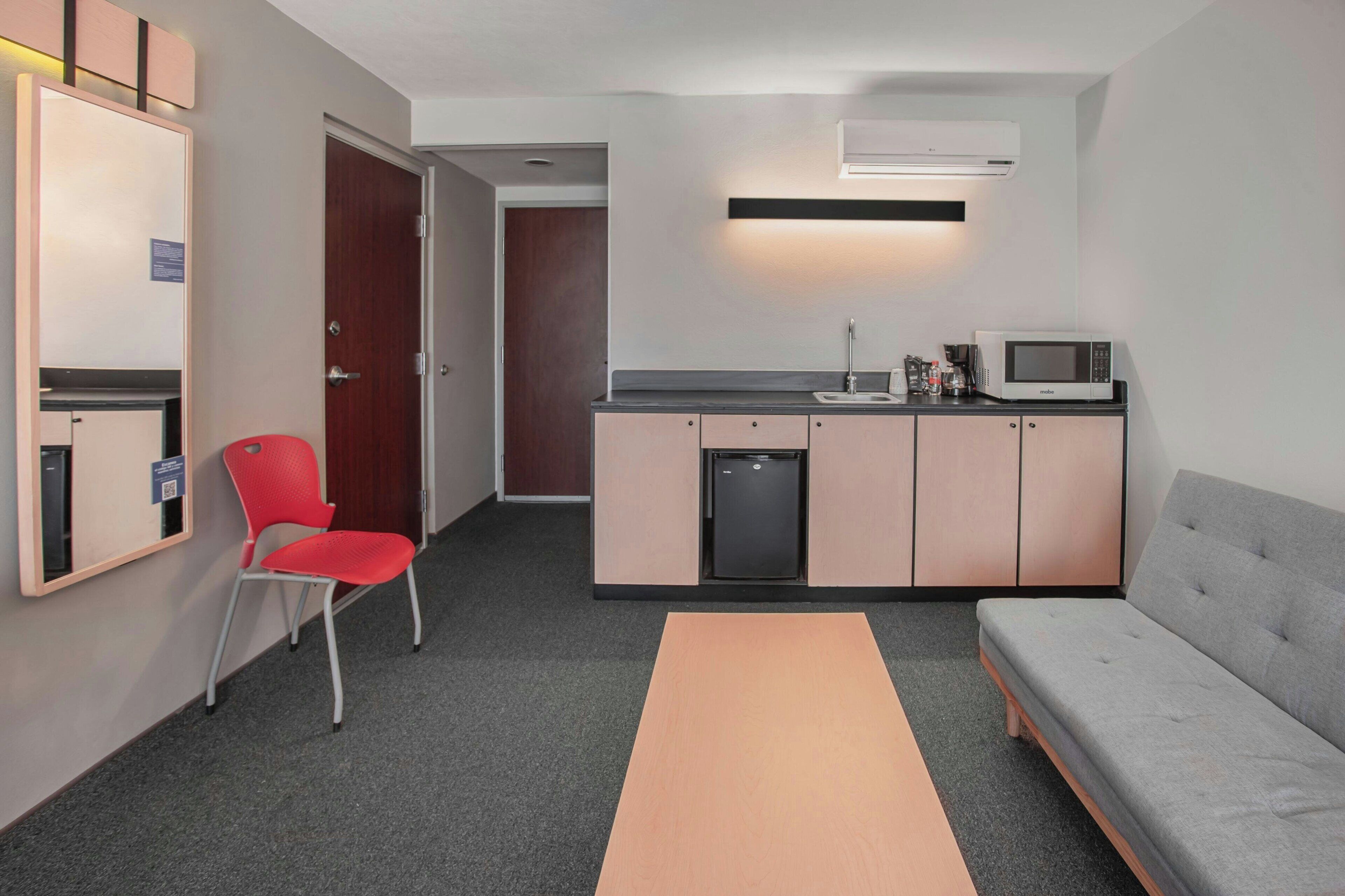 Una cocina compacta con microondas, lavamanos y un refrigerador pequeño. Hay una silla roja y un sofá gris en la habitación.