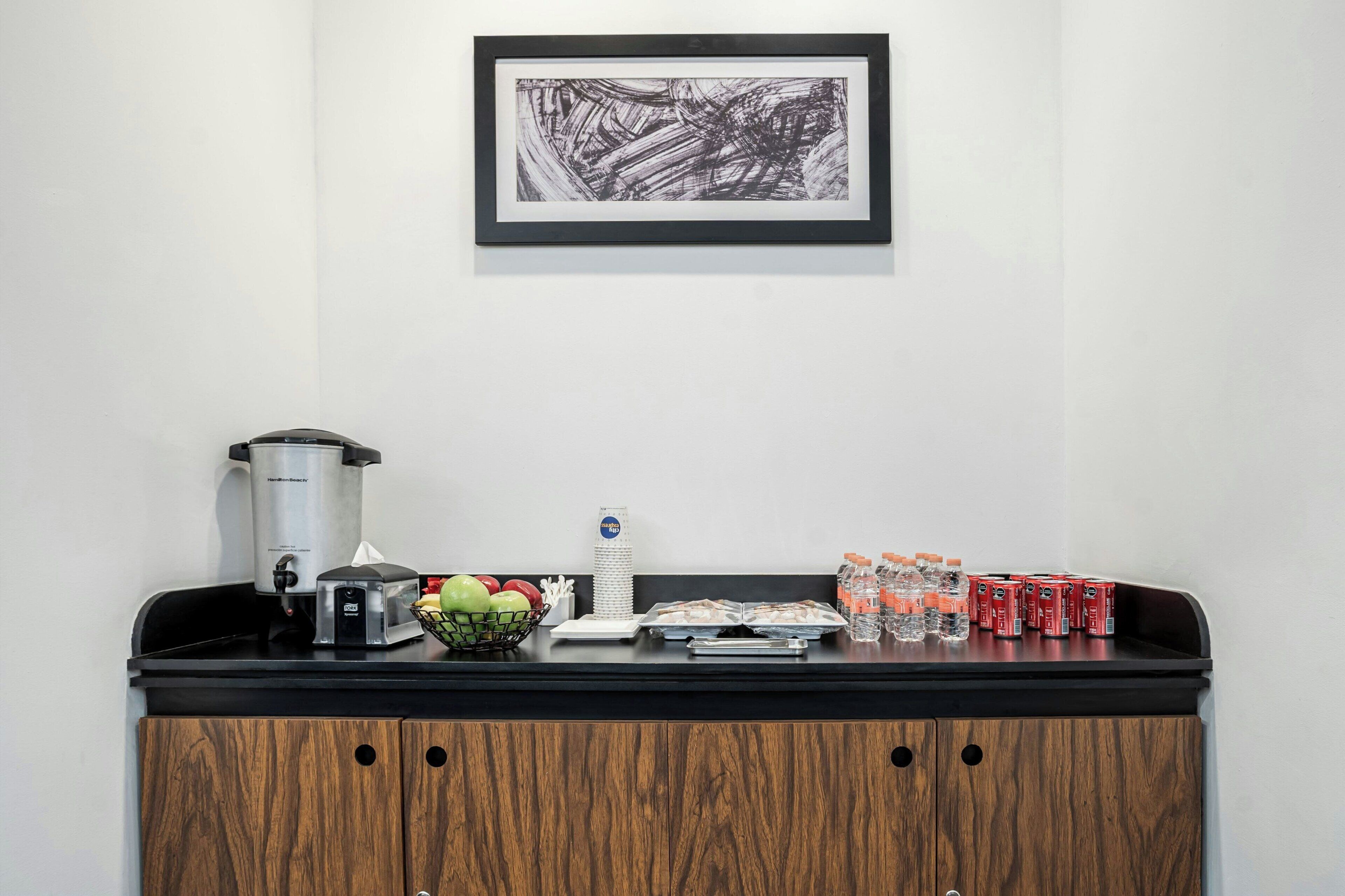 Una kitchenette con cafetera, tostadora y frutero. También hay agua embotellada y bebidas enlatadas sobre la encimera.