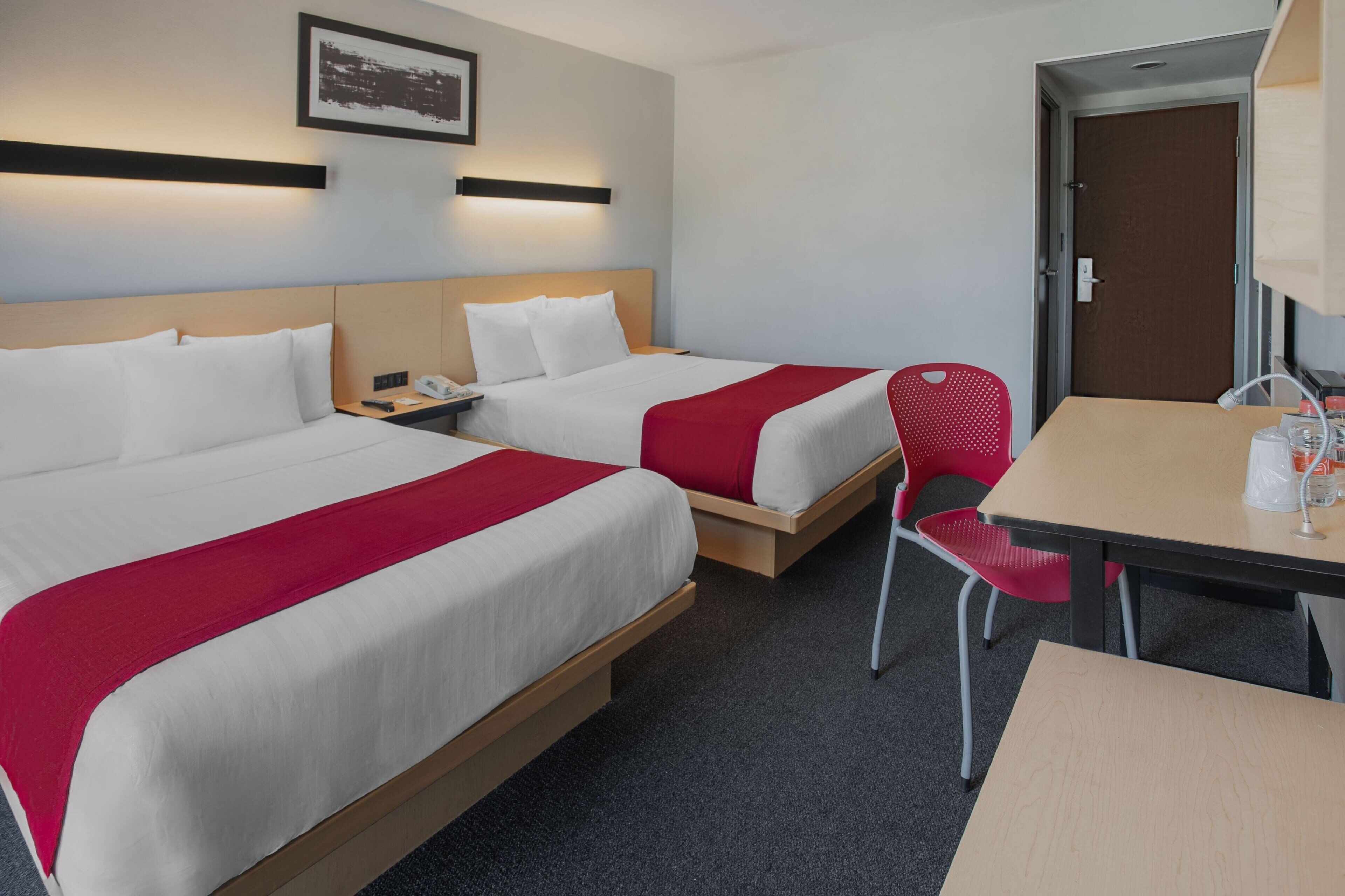 A hotel room with two beds, a desk, and a chair. There is a door and a framed picture on the wall.