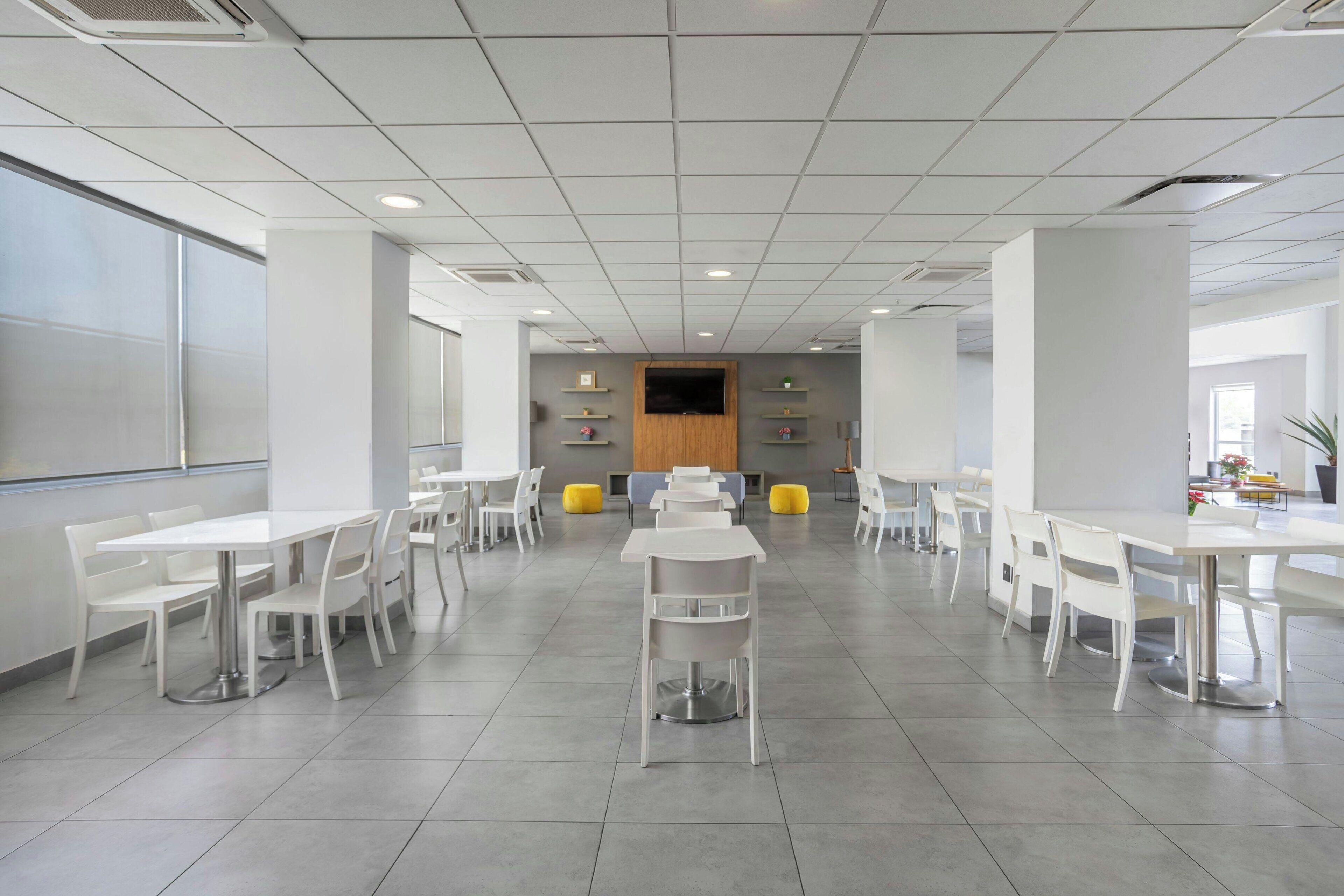 A spacious, modern dining area with white tables and chairs, a central seating area with a wooden panel, and a large window providing natural light.