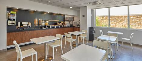 A modern cafe with white tables and chairs, a coffee machine, and a view of a brick wall through large windows.