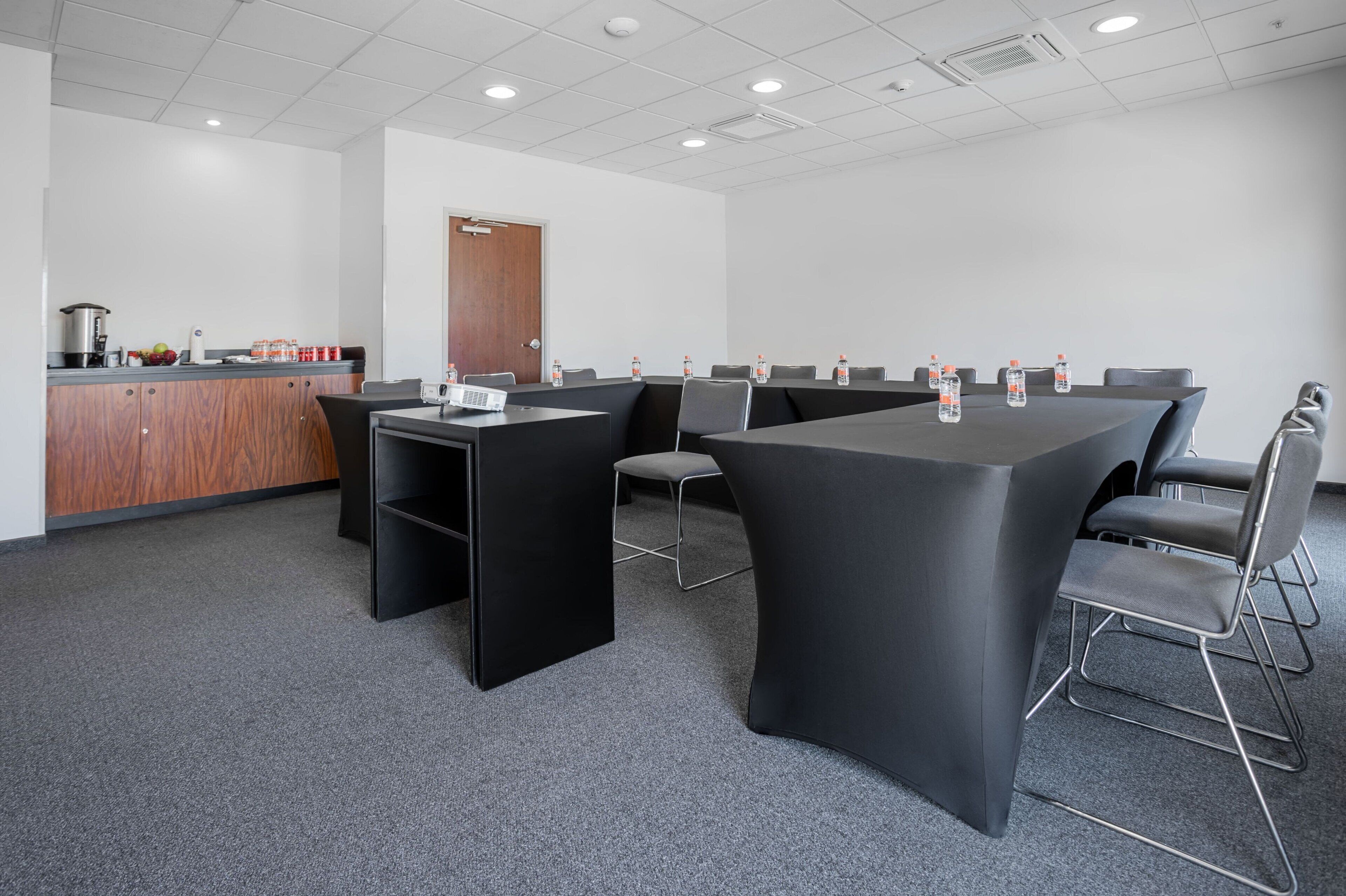 Un salón de conferencias con una mesa larga, sillas, una pequeña cocina y una puerta.