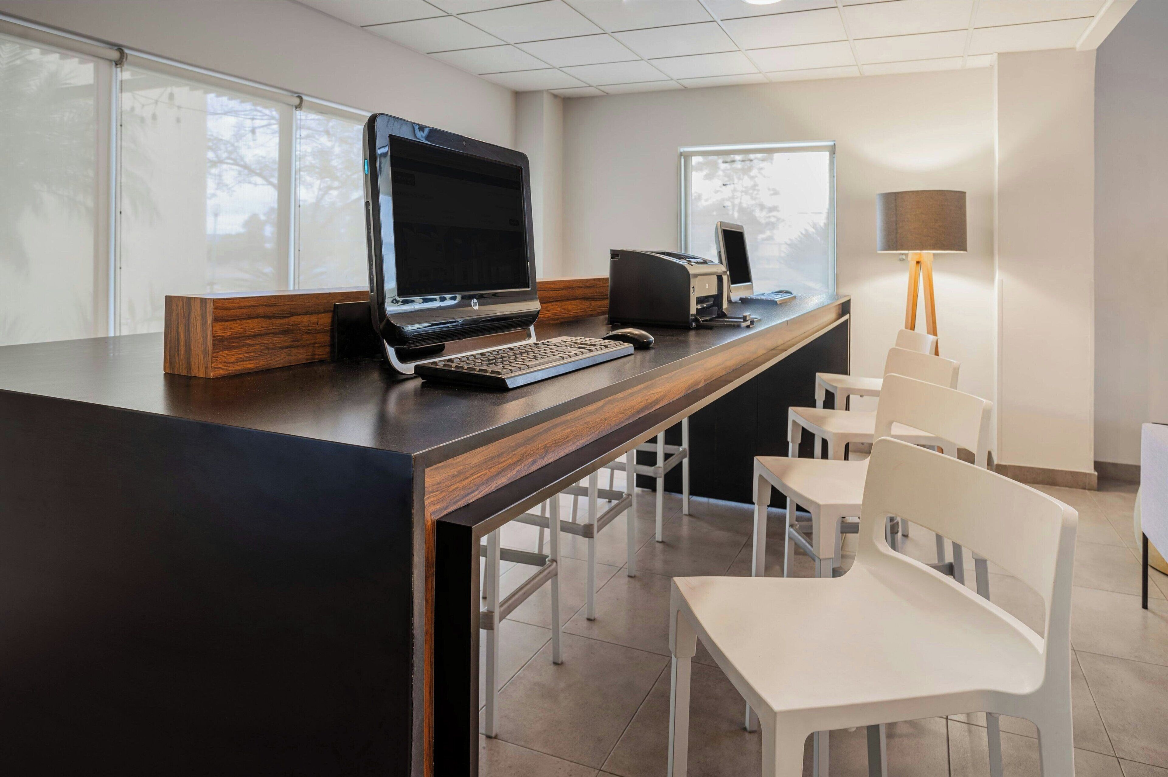 Un espacio de oficina moderno con un escritorio de madera largo, un ordenador de escritorio, una impresora y varias sillas blancas.