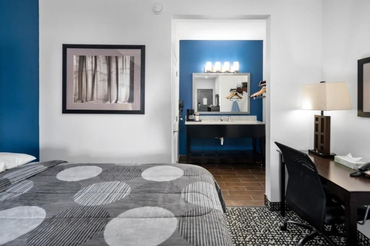 A hotel room with a bed, a desk, a chair, a desk lamp, a phone, a mirror, and a bathroom with a sink and vanity.