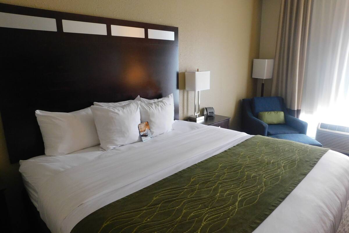 A hotel room with a large bed, a headboard, a desk with a lamp, a chair, and a window with curtains.