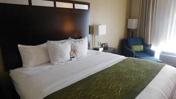 Ein Hotelzimmer mit einem großen Bett, einem Kopfteil, einem Schreibtisch mit Lampe, einem Sessel und einem Fenster mit Vorhängen.