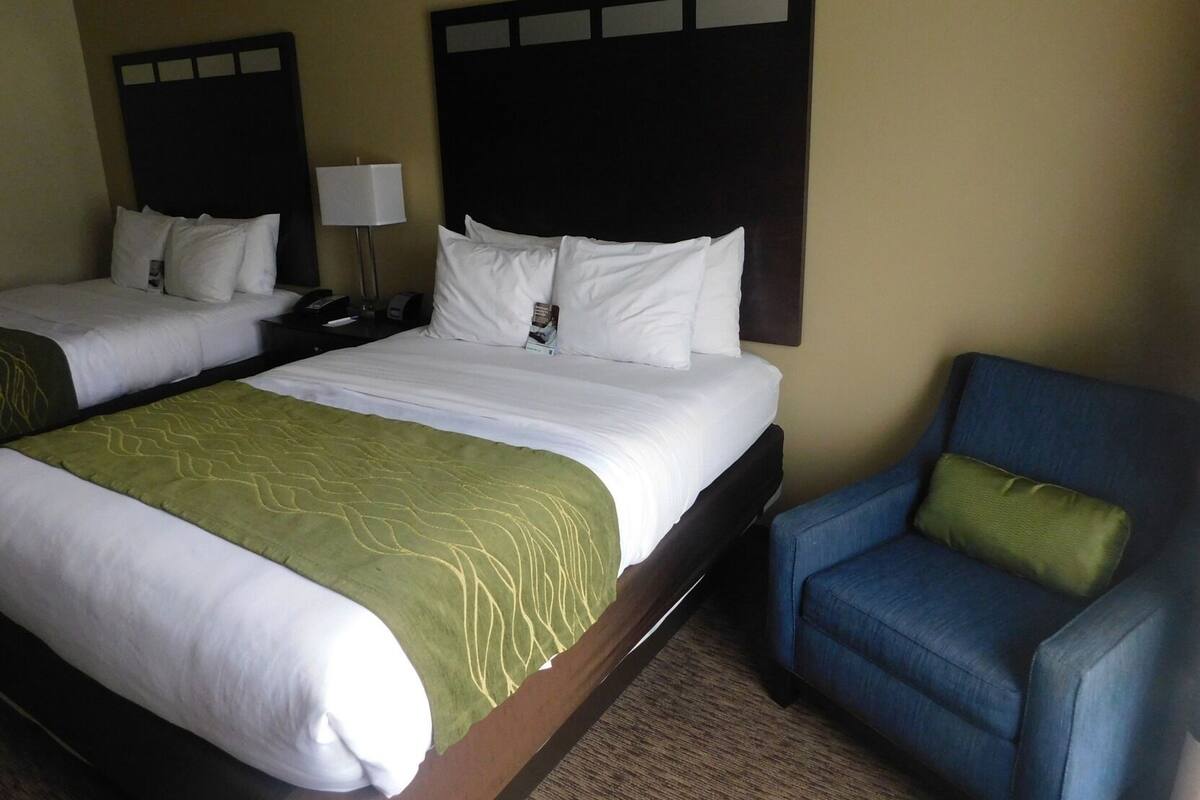 A hotel room with two beds, a desk, a chair, and a lamp.