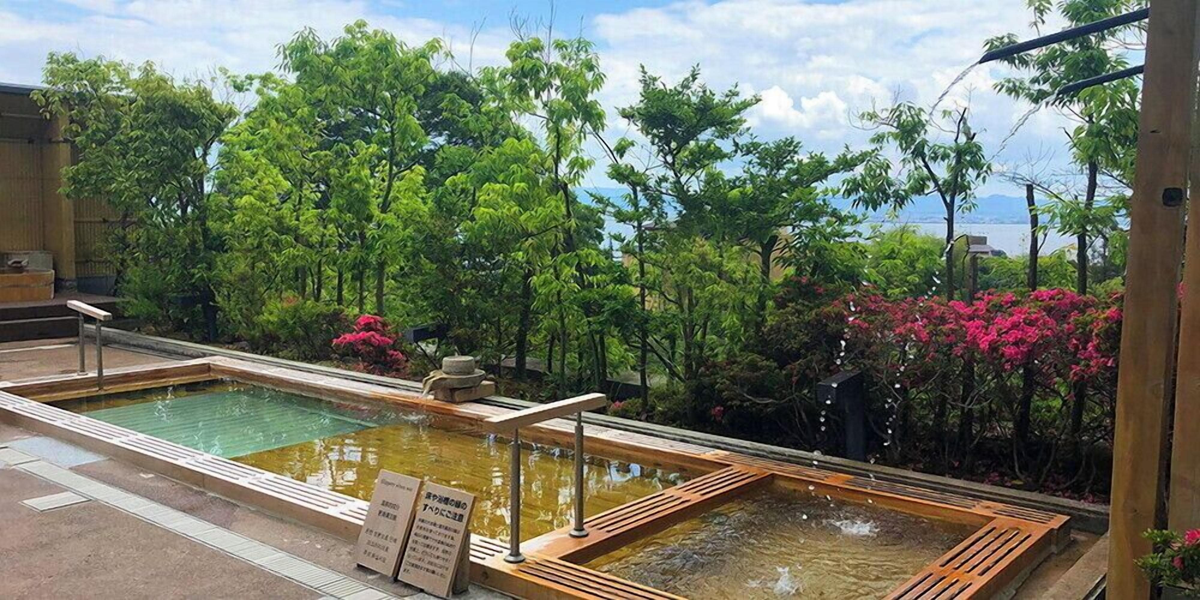 Un onsen japonais traditionnel, avec des bains en bois, offrant une vue sur une végétation luxuriante et un plan d’eau.