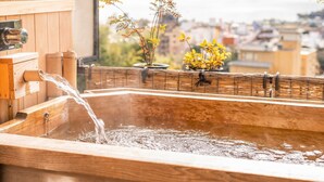 A wooden hot tub with water flowing, a window with a view of a cityscape and mountains, and two potted plants on the windowsill. - Satoyu Mukashibanashi Yuzanso Ryokan (Otsu)