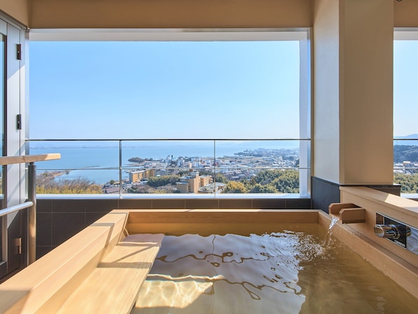 A modern bathroom with a large bathtub, a view of a cityscape, and a balcony with a railing. - Satoyu Mukashibanashi Yuzanso Ryokan (Otsu)
