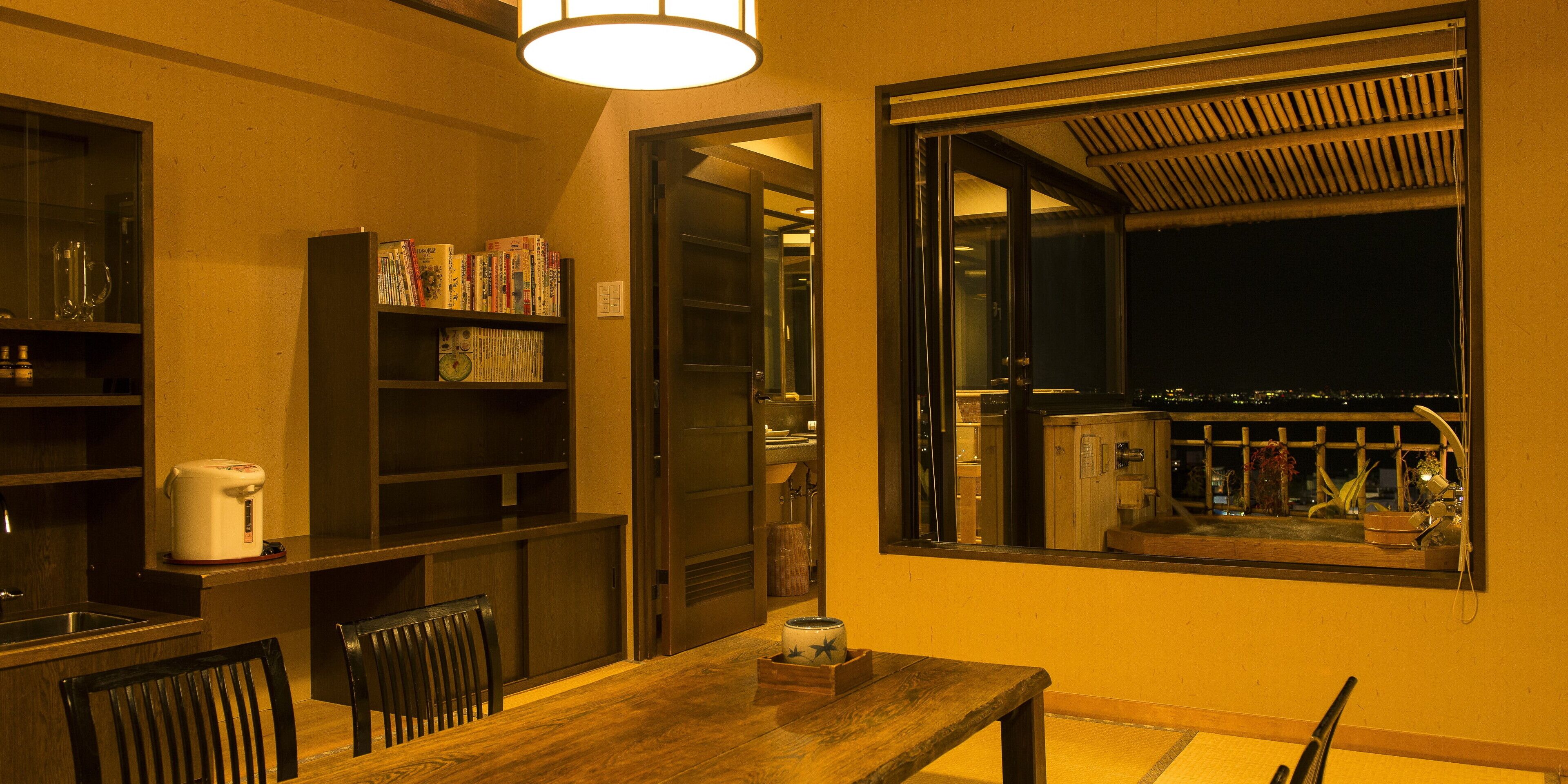 A dining area with a wooden table and chairs, a bookshelf with various books, a coffee maker, and a window with a view of the city at night.