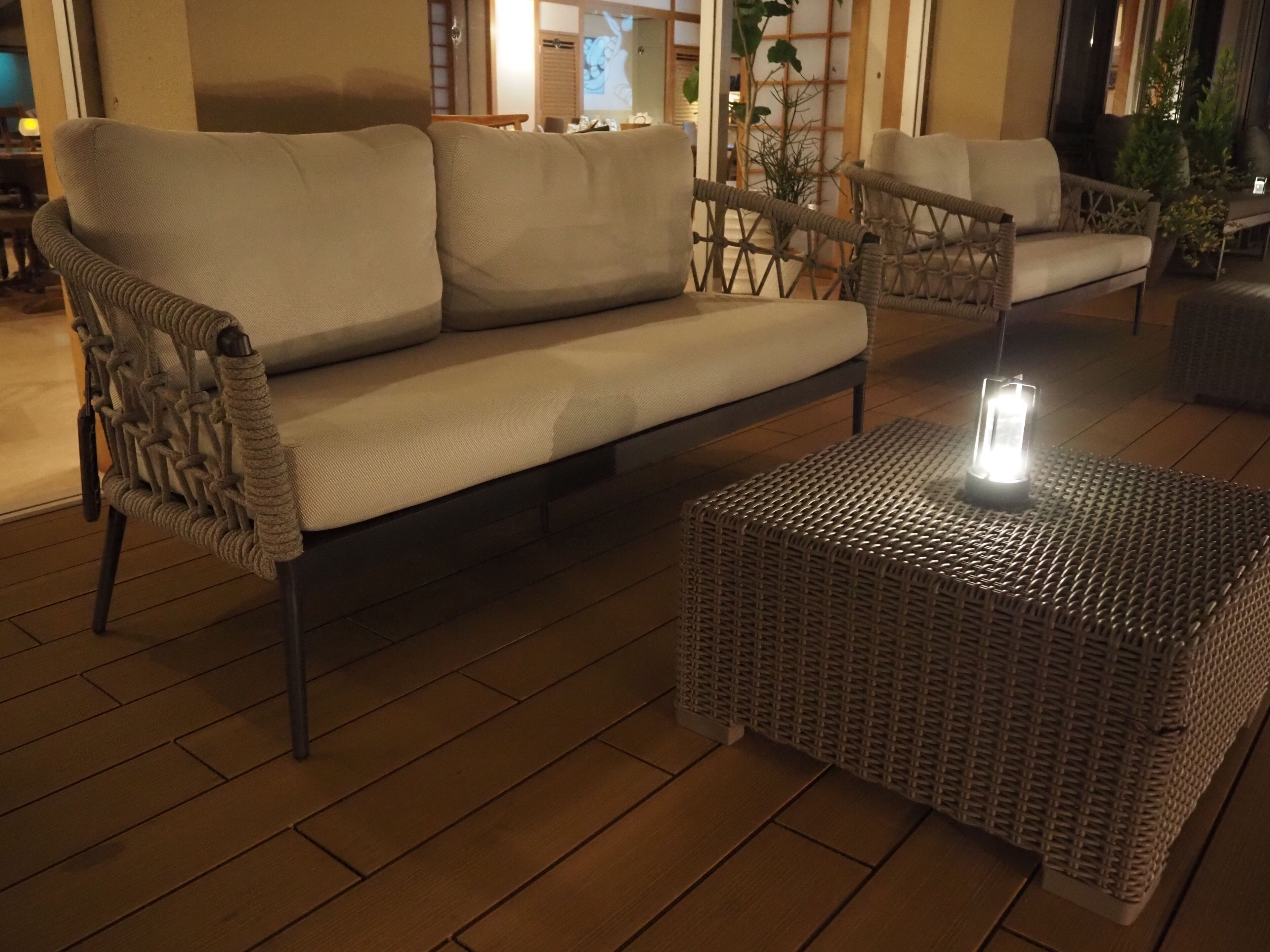 Un juego de sofá y sillón de mimbre sobre una terraza de madera, con un farol en una mesa tejida.