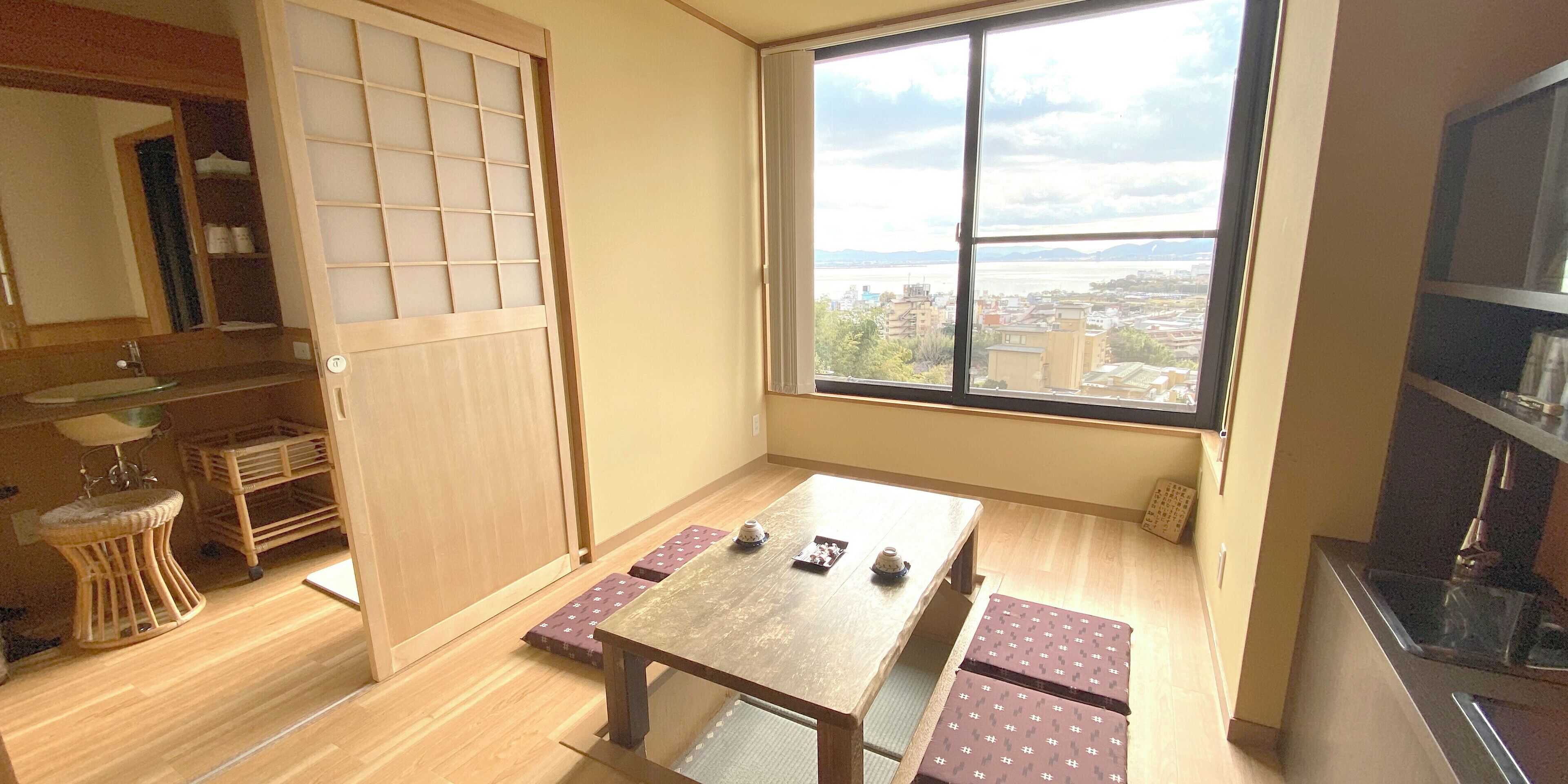 A room with a wooden floor, a tatami mat table, a low table, a window with a view of buildings and water, and a sliding door.