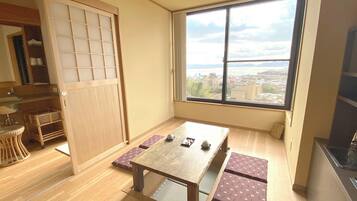 A room with a wooden floor, a tatami mat table, a low table, a window with a view of buildings and water, and a sliding door.