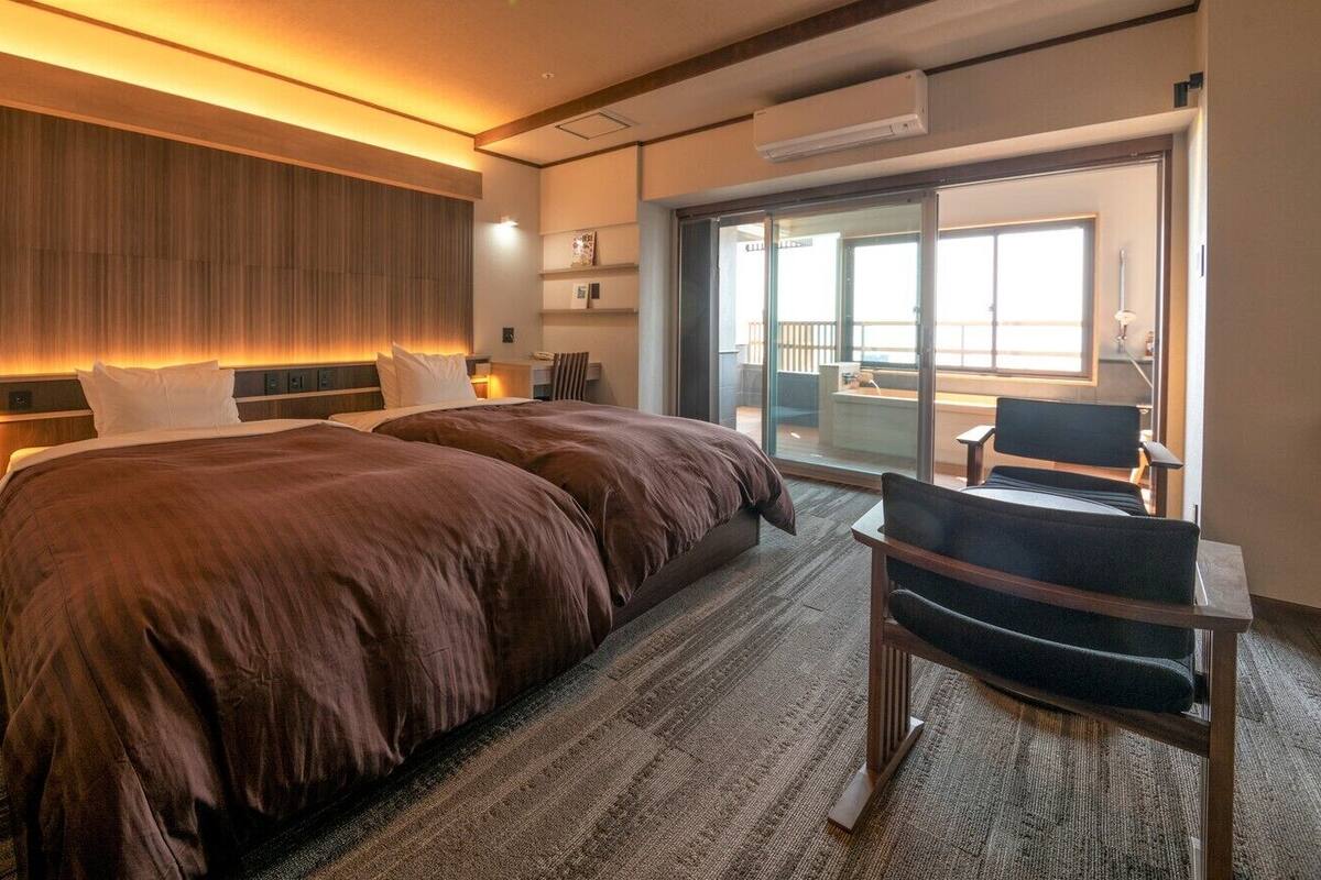 A hotel room with a large bed, a desk with a chair, a sliding glass door to a balcony, and a built-in shelf with decorative items.