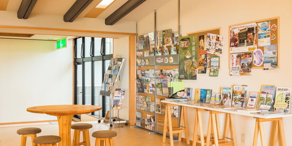 Een kamer met een houten tafel en krukken, een prikbord met verschillende spullen erop, en een raam met jaloezieën.