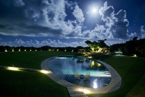 Piscina noturna com duas cadeiras, cercada por caminhos bem iluminados e árvores.