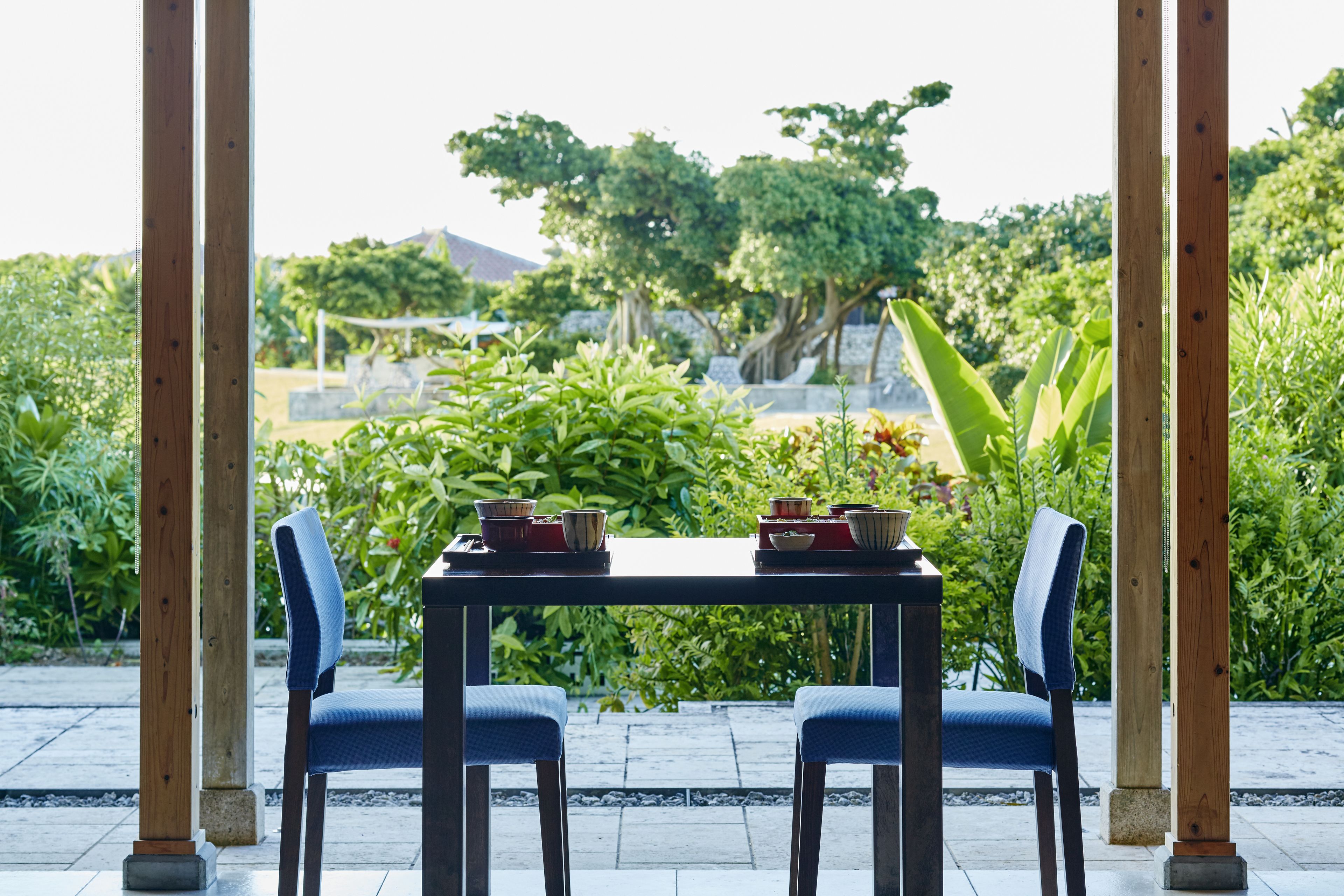 Una mesa dispuesta para dos personas, con sillas, rodeada de abundante follaje y vistas a árboles y plantas.